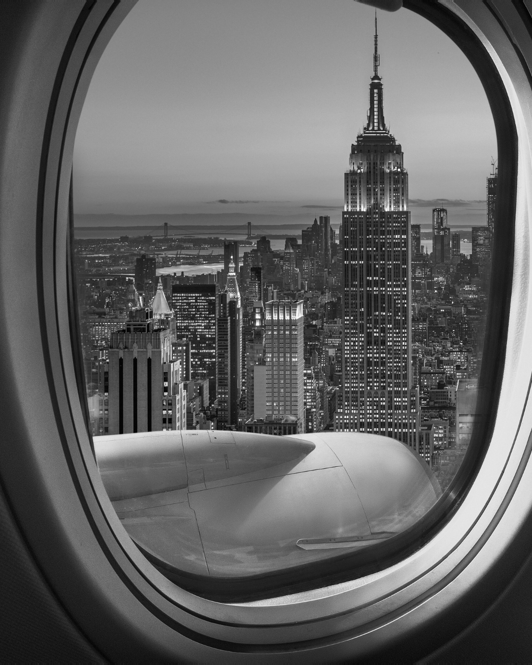 A black and white image of New York City featuring the Empire State Building, taken from a plane's window 