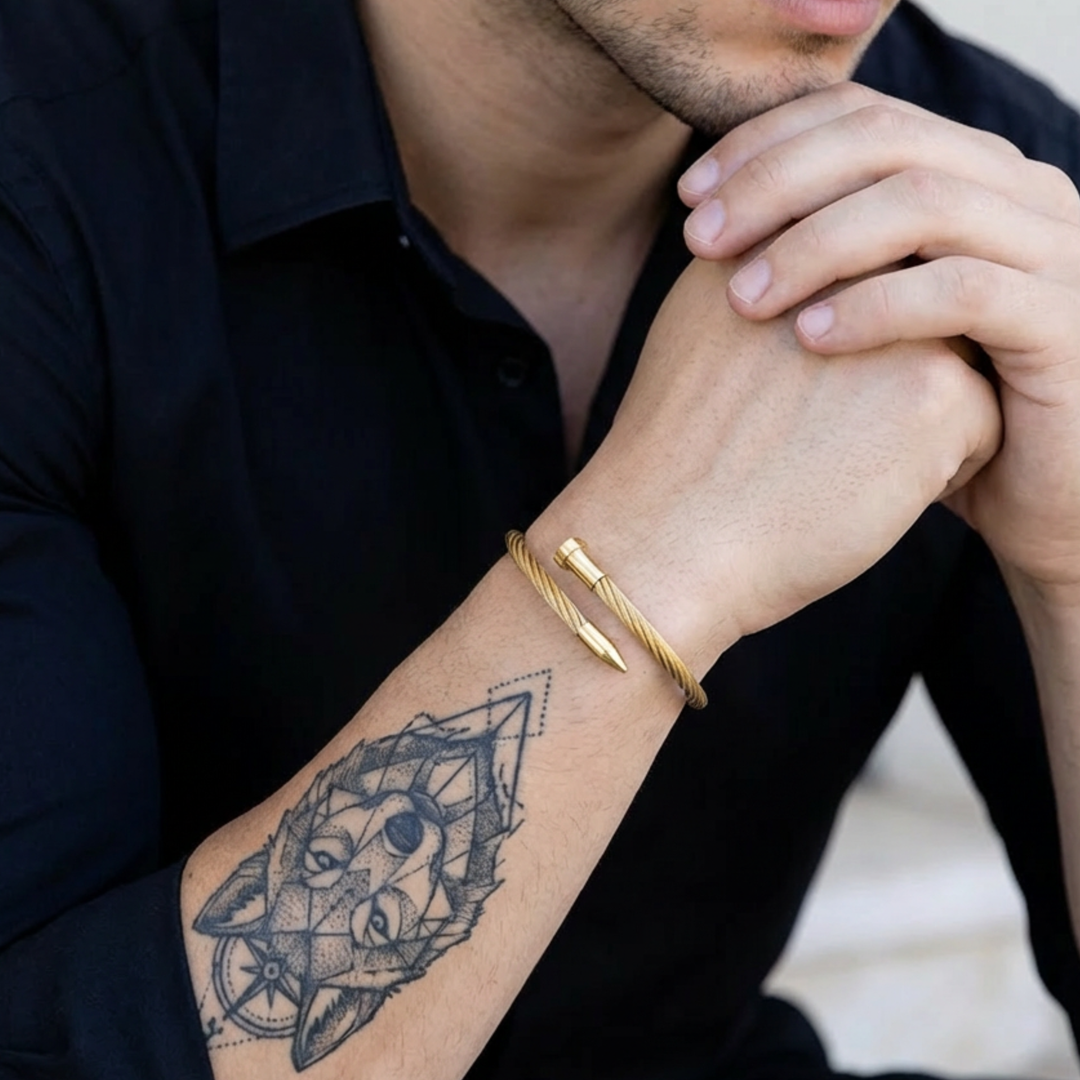 Person wearing a gold bracelet with a tattoo on their arm, sitting against a neutral background.