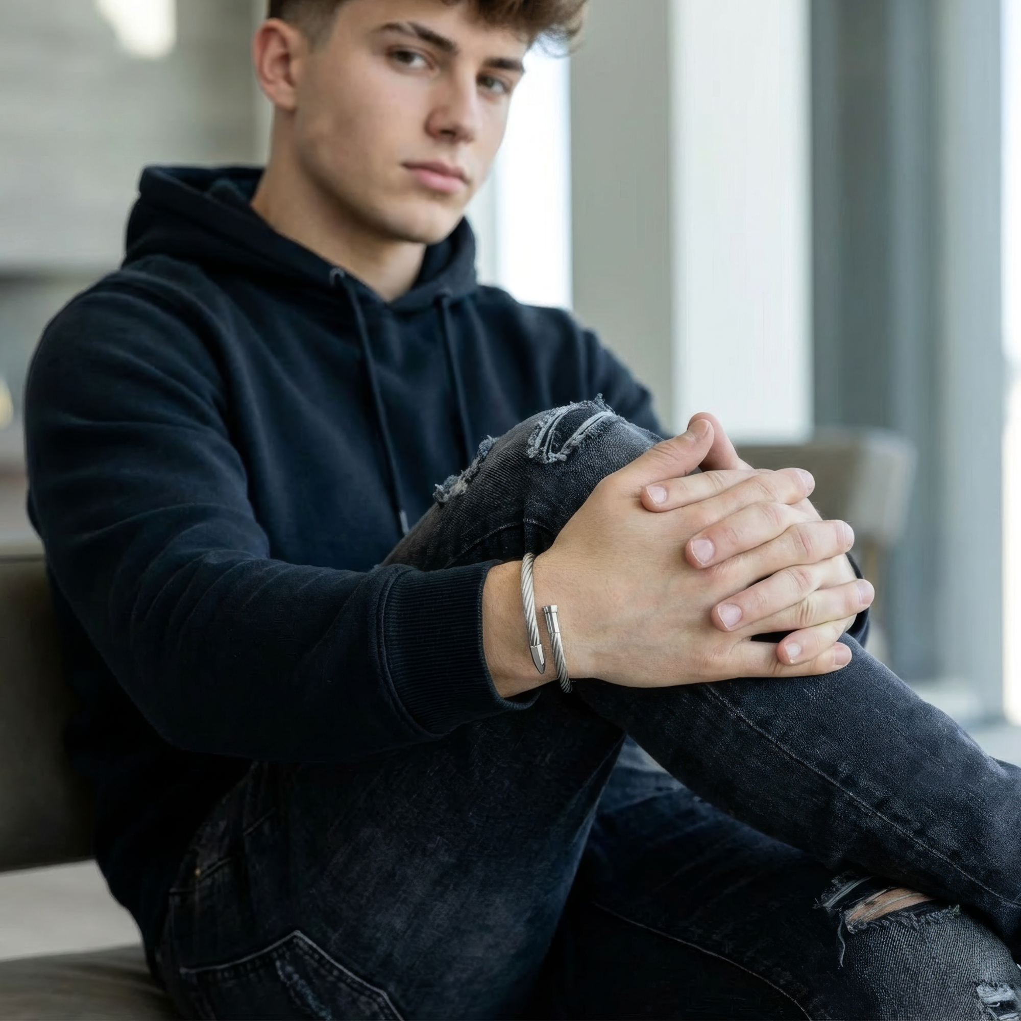 Person wearing a black hoodie and jeans sitting with hands clasped, a stainless steel bracelet on the their right wrist, looking off to the side.