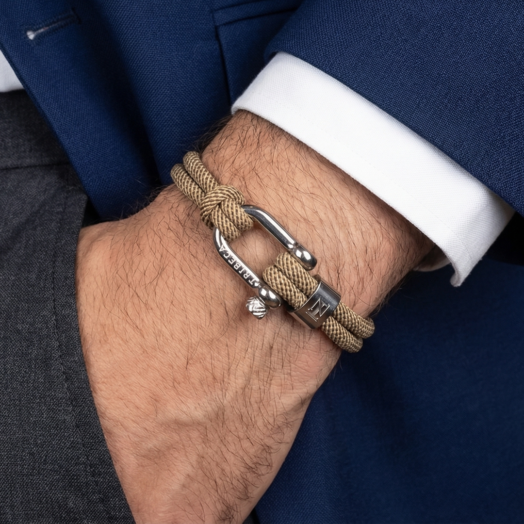 A man wearing a blue blazer and dress slacks with a beige rope bracelet with a metal clasp on his wrist. 